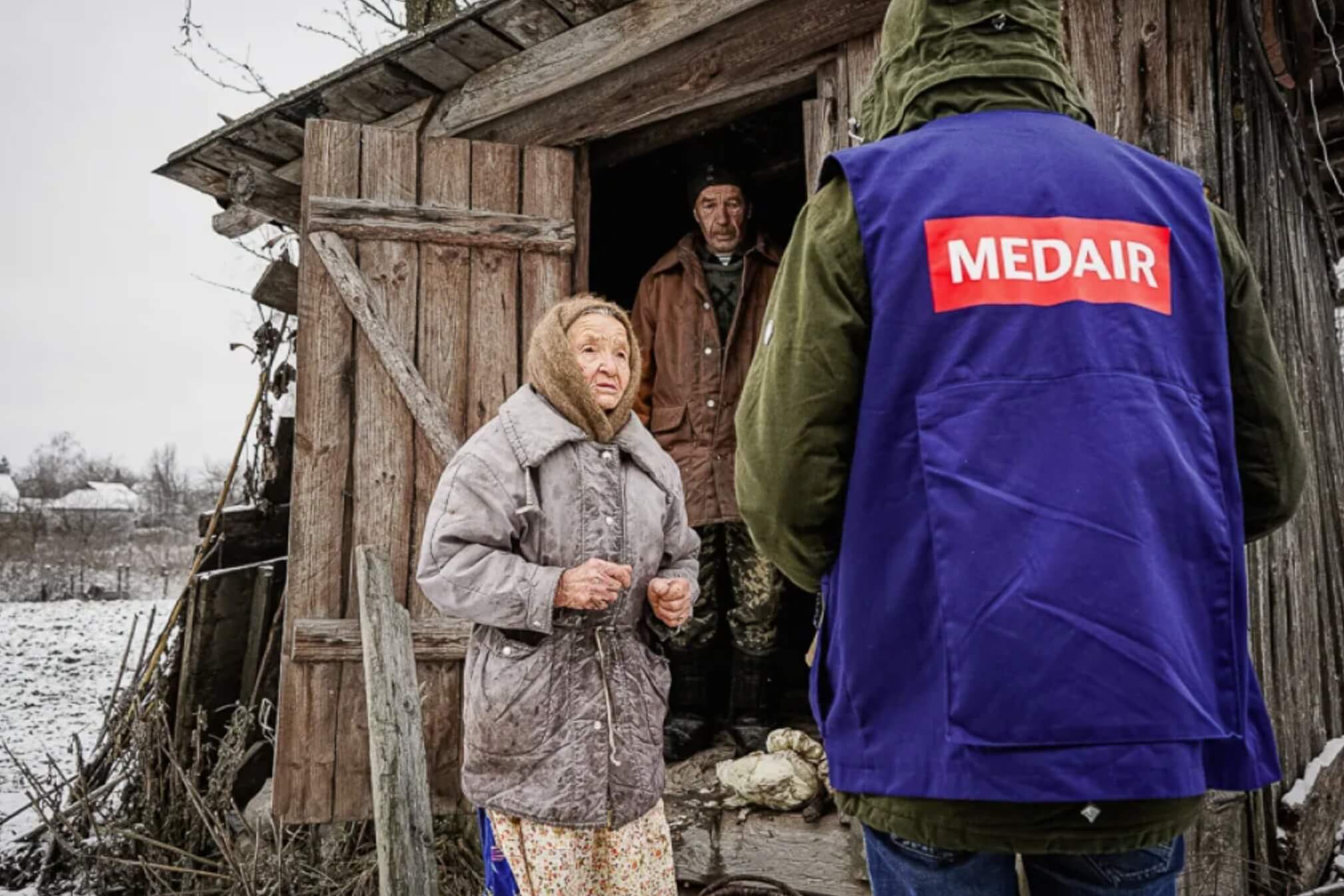 Une personne de dos avec un chasuble MEDAIR discutant avec une vieille dame devant une habitation en bois dans un paysage enneigé